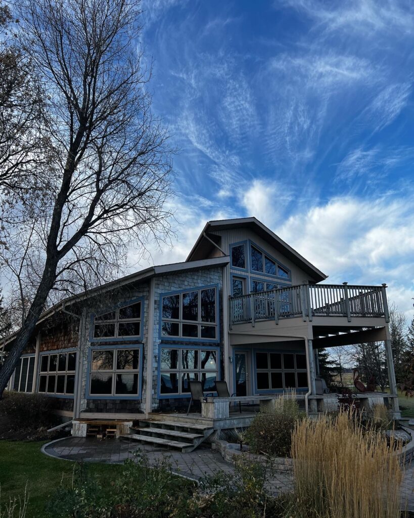 A two-story house with large, freshly cleaned windows and a balcony, surrounded by bare trees and a blue sky with wispy clouds.